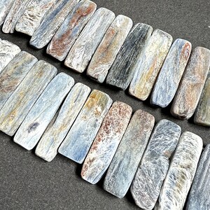 Pode incluir: Close-up de um fio de contas retangulares de cianita. As contas exibem uma mistura de tons azuis, cinzentos, castanhos e dourados, com varia&ccedil;&otilde;es naturais de cor e textura. As contas est&atilde;o dispostas em uma fileira, mostrando suas superf&iacute;cies polidas.
