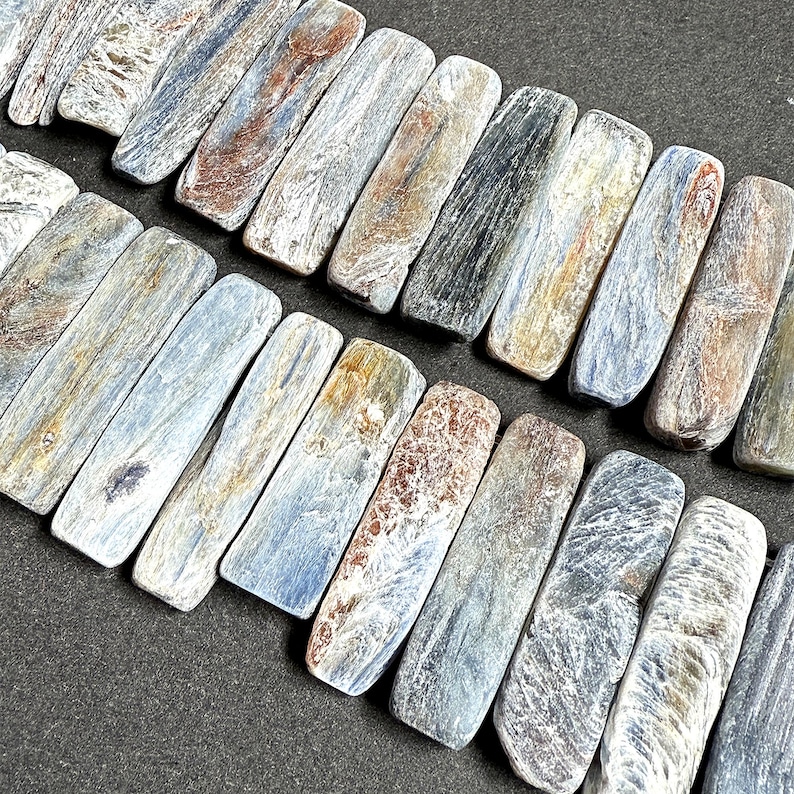 Pode incluir: Close-up de um fio de contas retangulares de cianita. As contas exibem uma mistura de tons azuis, cinzentos, castanhos e dourados, com varia&ccedil;&otilde;es naturais de cor e textura. As contas est&atilde;o dispostas em uma fileira, mostrando suas superf&iacute;cies polidas.