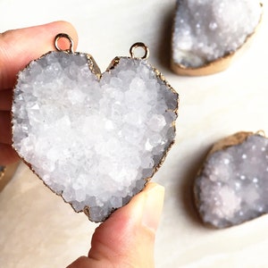 May include: A white heart-shaped crystal with a gold-toned metal rim. The crystal is made of many small, white crystals that are clustered together. The heart is being held by a hand.
