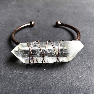 May include: A clear quartz crystal wrapped in copper wire, forming a bracelet. The crystal is a long, pointed shape, and the wire is wrapped around it in a criss-cross pattern.