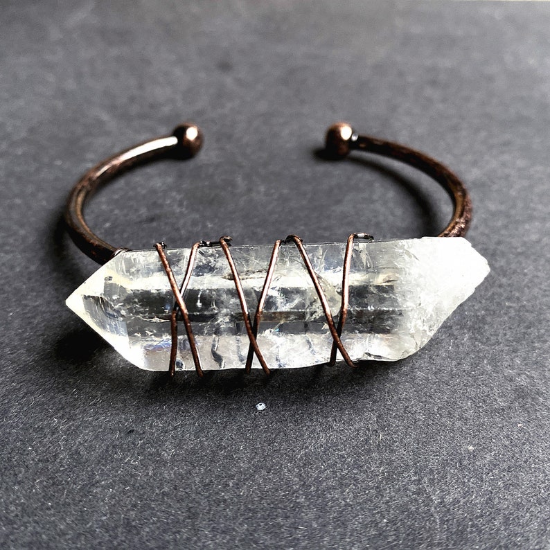 May include: A clear quartz crystal wrapped in copper wire, forming a bracelet. The crystal is a long, pointed shape, and the wire is wrapped around it in a criss-cross pattern.