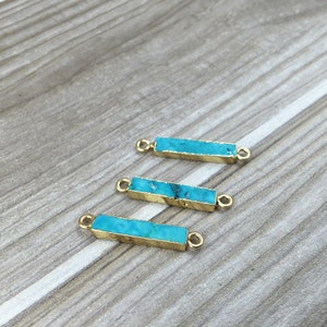 Puede incluir: Tres conectores de barra rectangulares de color azul turquesa con bucles de metal dorado en cada extremo. Los conectores están dispuestos en forma escalonada sobre una superficie de madera.