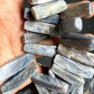 Pode incluir: Close-up de uma cole&ccedil;&atilde;o de contas de pedras preciosas de cianita azul-cinzento. As contas s&atilde;o retangulares com bordas arredondadas e mostram veios e varia&ccedil;&otilde;es de cor naturais. As contas s&atilde;o enfiadas.