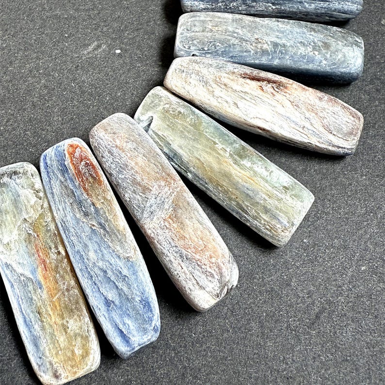 Pode incluir: Close-up de v&aacute;rias pedras retangulares polidas em tons de azul, cinza e castanho. As pedras t&ecirc;m uma apar&ecirc;ncia lisa e texturizada com varia&ccedil;&otilde;es naturais de cor e padr&atilde;o. As pedras est&atilde;o dispostas sobre uma superf&iacute;cie escura.