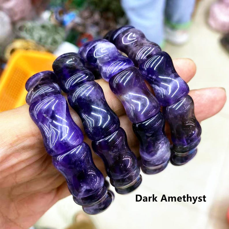 Op de afbeelding: Vier donkere amethistarmbanden, elk bestaande uit gesneden, cilindrische kralen. De kralen zijn diep paars met lichtere paarse adering. De armbanden worden in een hand gehouden, met de tekst "Dark Amethyst" zichtbaar.