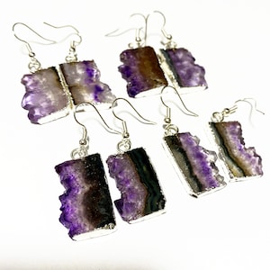 Puede incluir: Cuatro pares de pendientes de plata con detalles de piedra de amatista. Las piedras de amatista son de color morado intenso con una textura natural y rugosa. Los pendientes tienen forma rectangular con un respaldo de plata.