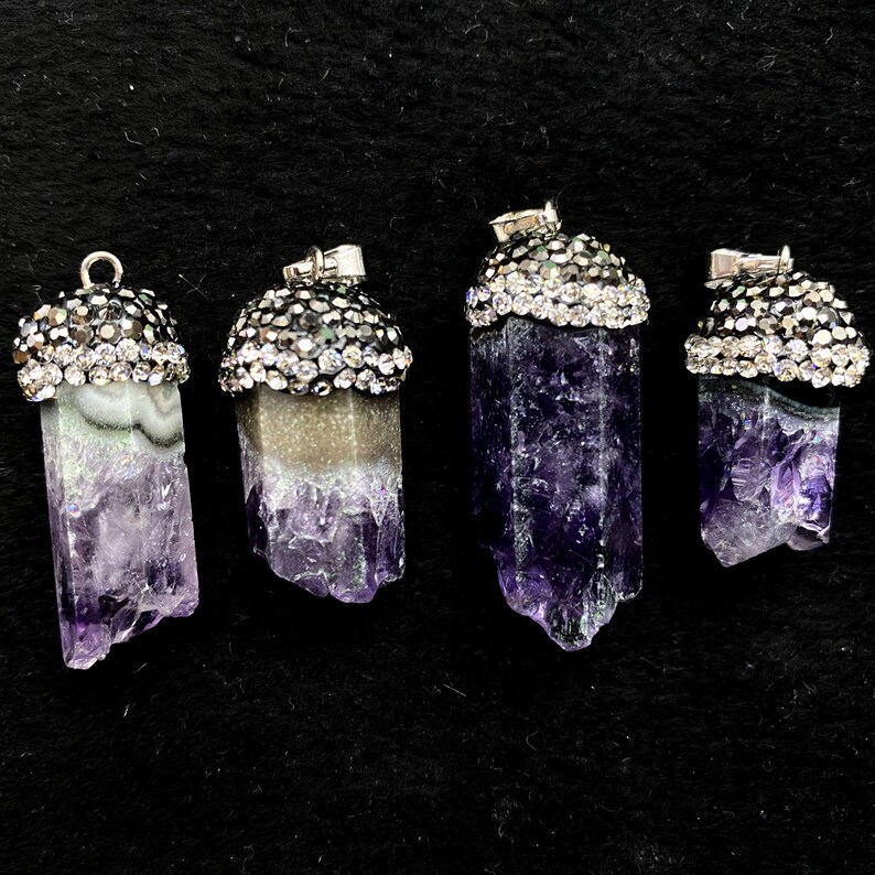 Puede incluir: Cuatro colgantes de cristal de amatista con engastes plateados y detalles de diamantes de imitaci&oacute;n transparentes. Los colgantes son de diferentes tama&ntilde;os y formas, y todos est&aacute;n colocados sobre un fondo negro.