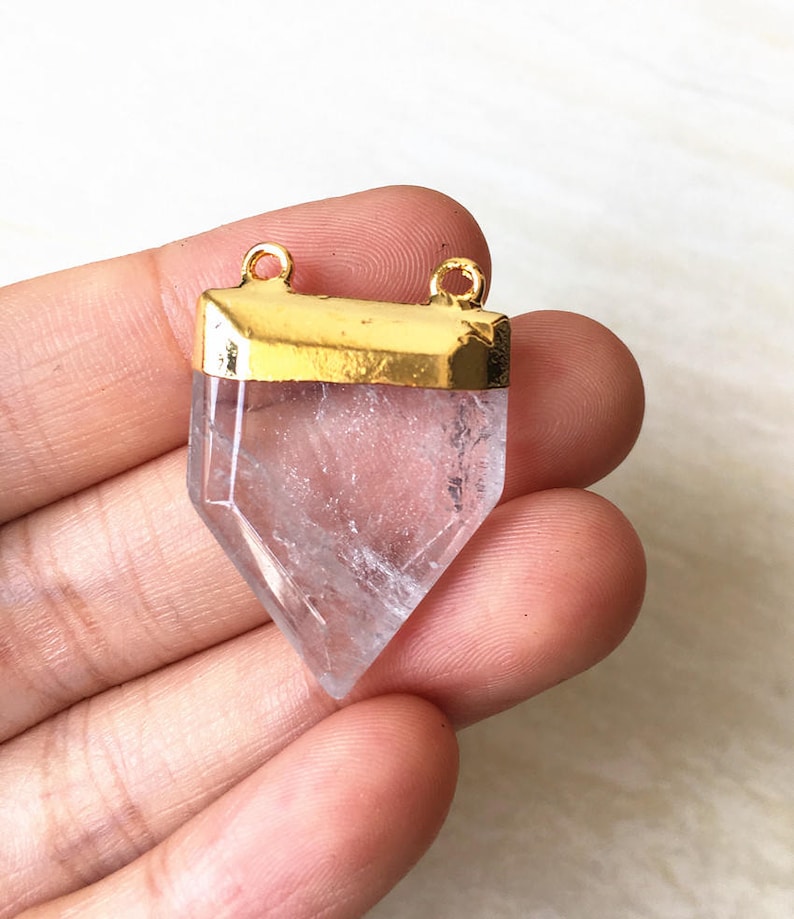 Puede incluir: Un colgante de cristal de cuarzo transparente con una parte superior chapada en oro y dos bucles para sujetar una cadena. El cristal tiene forma de punta y un dise&ntilde;o facetado.