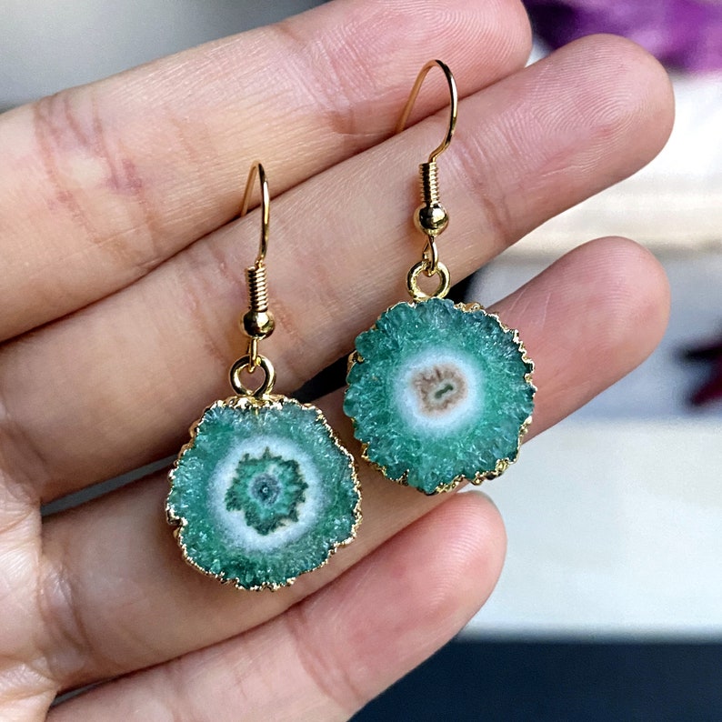 Puede incluir: Un par de pendientes de oro con detalles de piedra de &aacute;gata verde. Los pendientes presentan una piedra redonda verde con un centro blanco y un bucle de alambre dorado.