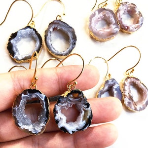May include: A pair of gold-toned earrings with agate stone accents. The earrings feature a round, hollowed-out agate stone with a white and gray interior and a black exterior. The agate stones are attached to gold-toned hooks.