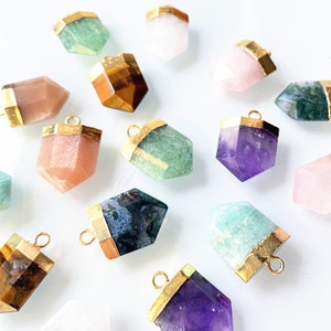 May include: Assortment of gold-rimmed crystal point pendants in various colours, including green, pink, purple, brown, and white. The pendants are arranged on a white surface.