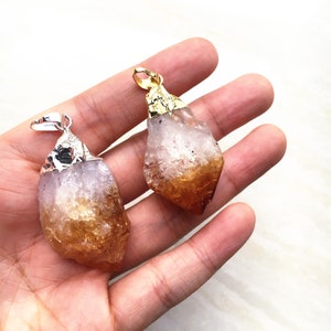 Puede incluir: Dos colgantes de cristal de citrino en bruto con detalles dorados y plateados. Los cristales son de color naranja claro con un centro transparente y translúcido.