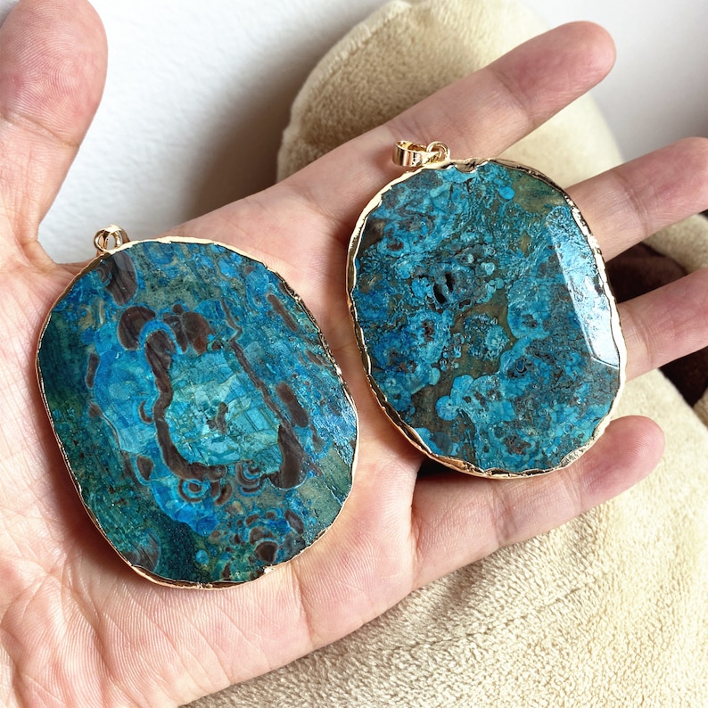 May include: Two oval-shaped turquoise and brown agate pendants with gold trim. The pendants are on a hand.