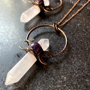 May include: A rose quartz crystal pendant necklace with a copper crescent moon design and an amethyst stone accent. The necklace is on a copper chain.