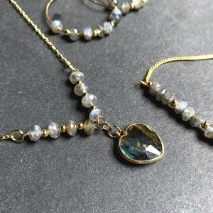 May include: A gold necklace with a heart-shaped pendant made of iridescent labradorite gemstone. The necklace is made with small round labradorite beads and a delicate gold chain.