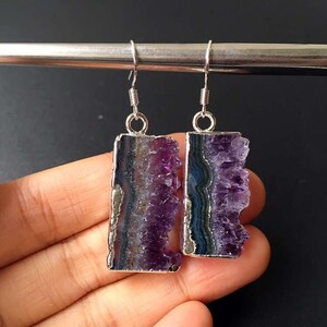 Puede incluir: Un par de pendientes de plata con gotas de cristal de amatista. Los cristales son de color morado oscuro y tienen una textura natural y rugosa. Los pendientes son una pieza llamativa y ser&iacute;an perfectos para una ocasi&oacute;n especial.