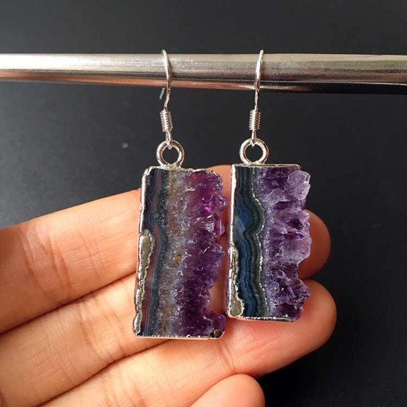 Puede incluir: Un par de pendientes de plata con gotas de cristal de amatista. Los cristales son de color morado oscuro y tienen una textura natural y rugosa. Los pendientes son una pieza llamativa y ser&iacute;an perfectos para una ocasi&oacute;n especial.