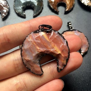 May include: A crescent moon shaped pendant made from a red, white, and brown stone with a black metal trim. The pendant is held in a hand.