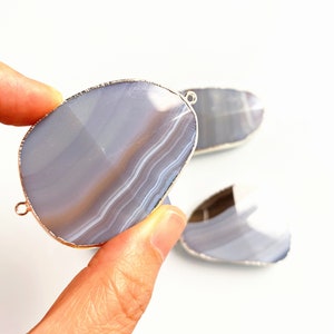 May include: Two silver-rimmed agate slices with a light blue and white pattern. The slices are oval-shaped and have a smooth, polished surface.