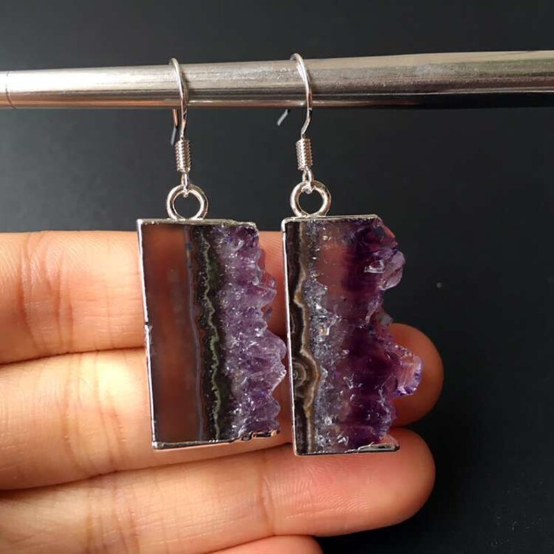 Puede incluir: Un par de pendientes de plata con gotas de cristal de amatista. Los cristales son de color morado oscuro y tienen una textura natural y rugosa. Los pendientes son un accesorio &uacute;nico y elegante.