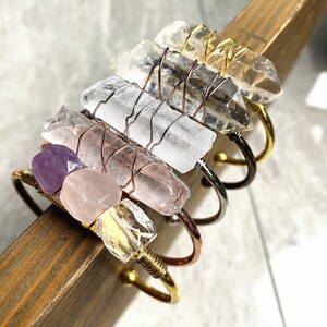 May include: A set of five crystal bracelets with wire wrapped around the crystals. The bracelets are made with clear quartz, rose quartz, amethyst, and citrine crystals. The bracelets are stacked on top of each other and are on a wooden surface.