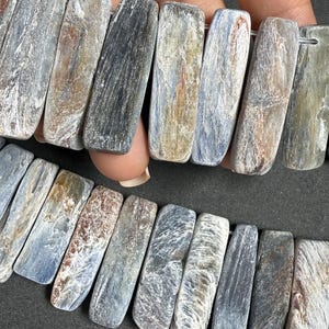 Pode incluir: Um fio de contas retangulares de cianita. As contas exibem uma mistura de tons azuis, cinzentos e castanhos com veios naturais. As pedras s&atilde;o enfiadas, mostrando suas superf&iacute;cies polidas e padr&otilde;es &uacute;nicos. Ideal para fazer joias.