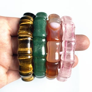 Op de afbeelding: Vier edelsteenarmbanden in bruin, groen, oranje en roze tinten. Elke armband heeft een bamboe-achtig ontwerp met cilindrische kralen. De armbanden worden in een hand tegen een witte achtergrond gehouden.