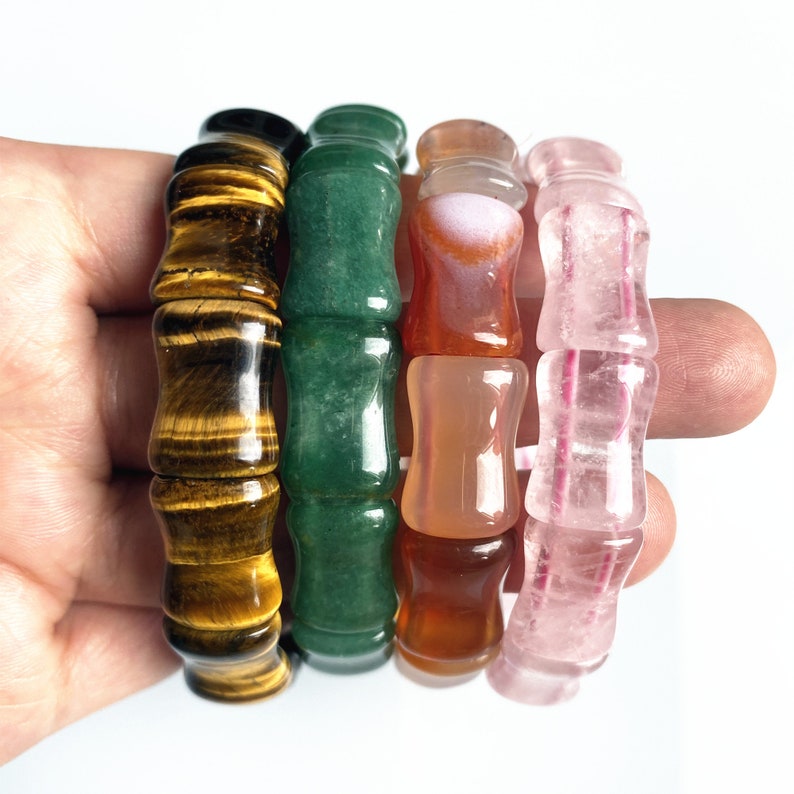 Op de afbeelding: Vier edelsteenarmbanden in bruin, groen, oranje en roze tinten. Elke armband heeft een bamboe-achtig ontwerp met cilindrische kralen. De armbanden worden in een hand tegen een witte achtergrond gehouden.