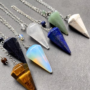 May include: Seven crystal pendulums on silver chains, each with a different color and shape. The crystals are clear quartz, blue lapis lazuli, green aventurine, white howlite, opal, black obsidian, and tiger's eye.