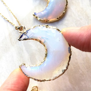 May include: A gold-rimmed crescent moon pendant made of white opal. The pendant is on a gold chain and is being held by two fingers.