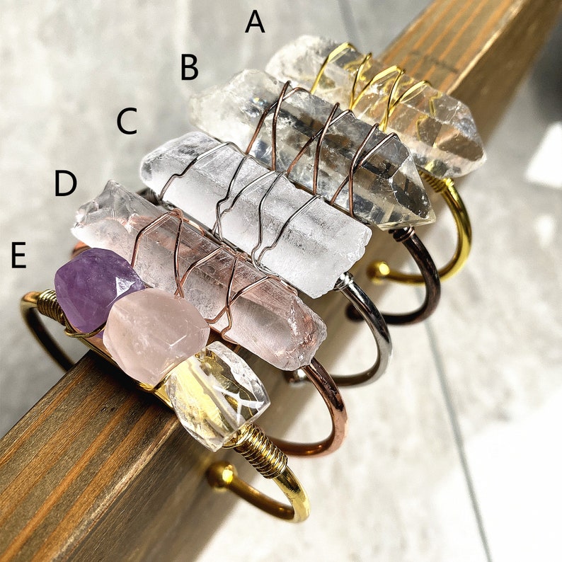 May include: Five crystal bracelets with different colored stones and gold wire wrapped around them. The bracelets are stacked on top of each other, with the top bracelet featuring a clear crystal, a pink crystal, and a purple crystal.
