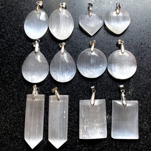 May include: Twelve selenite crystal pendants with silver bail settings. The pendants are in various shapes, including oval, round, heart, teardrop, and rectangular. The crystals are translucent and have a white, milky colour.