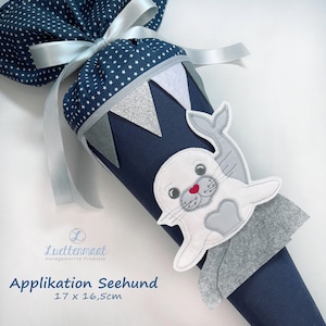 Könnte beinhalten: Marineblauer Schultüte mit weißem Sternmuster und silbernem Band. Die Tüte hat eine Applikation eines weißen Seehunds mit roter Nase und grauen Akzenten. Der Text "Applikation Seehund" ist sichtbar. Maße: 17 x 16,5 cm.