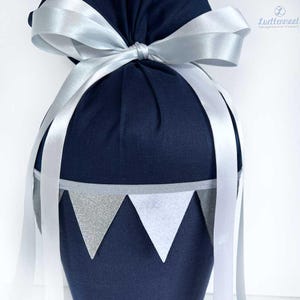 May include: A navy blue fabric gift bag with a white satin ribbon tied in a bow. The bag has a silver and white bunting trim around the top.