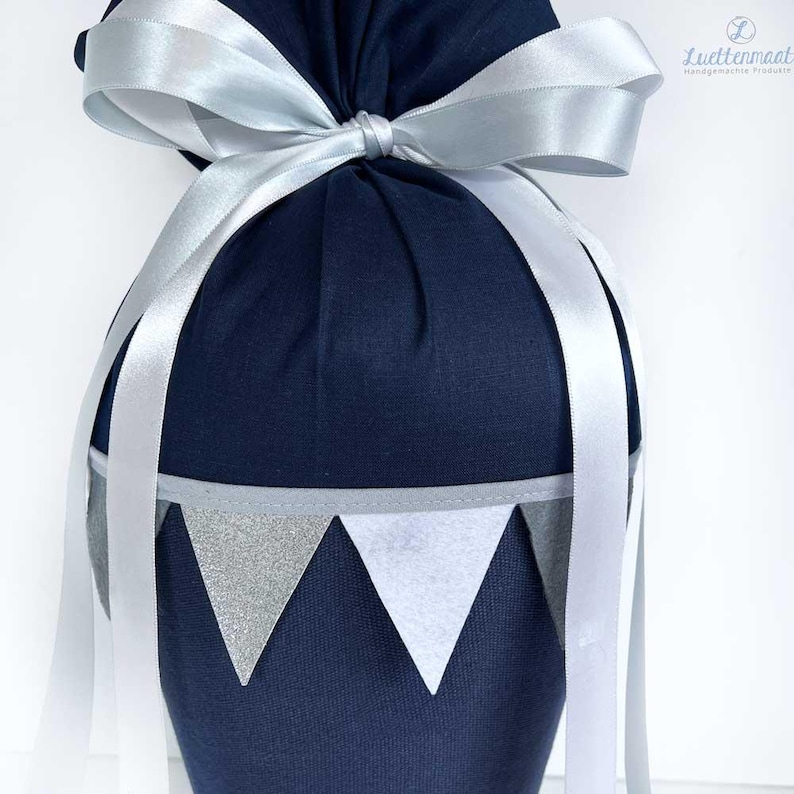 May include: A navy blue fabric gift bag with a white satin ribbon tied in a bow. The bag has a silver and white bunting trim around the top.