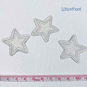 May include: Three silver glitter star-shaped iron-on patches, each with a white outline, on a white background. A measuring tape is visible at the bottom of the image.