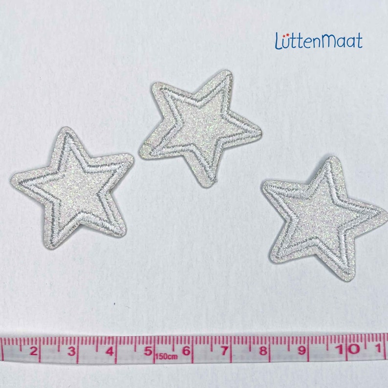 May include: Three silver glitter star-shaped iron-on patches, each with a white outline, on a white background. A measuring tape is visible at the bottom of the image.