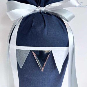 May include: A navy blue fabric gift bag with a white ribbon and silver triangle accents. The bag is tied with a bow and has a white trim around the top.