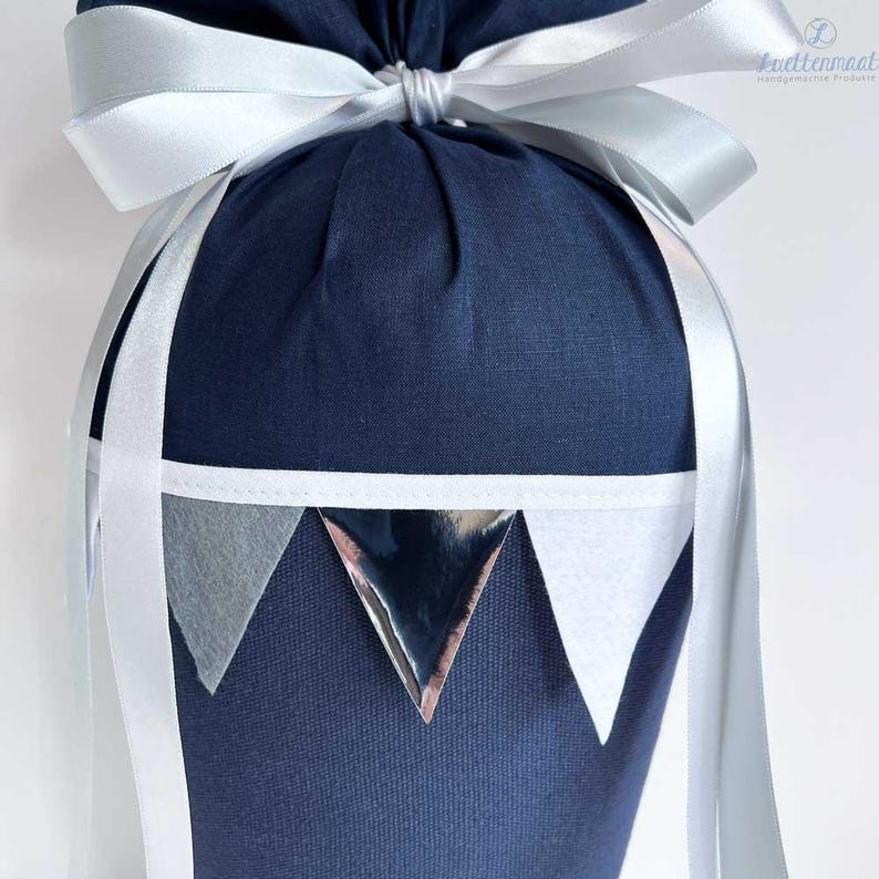 May include: A navy blue fabric gift bag with a white ribbon and silver triangle accents. The bag is tied with a bow and has a white trim around the top.