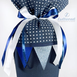 May include: A blue and white fabric gift bag with a white star pattern. The bag is tied with a white ribbon and blue ribbon. The bag has a blue and white fabric trim and a blue and silver glitter fabric trim. The bag has a tag that says "Luetjenmaat Handgemachte Produkte".