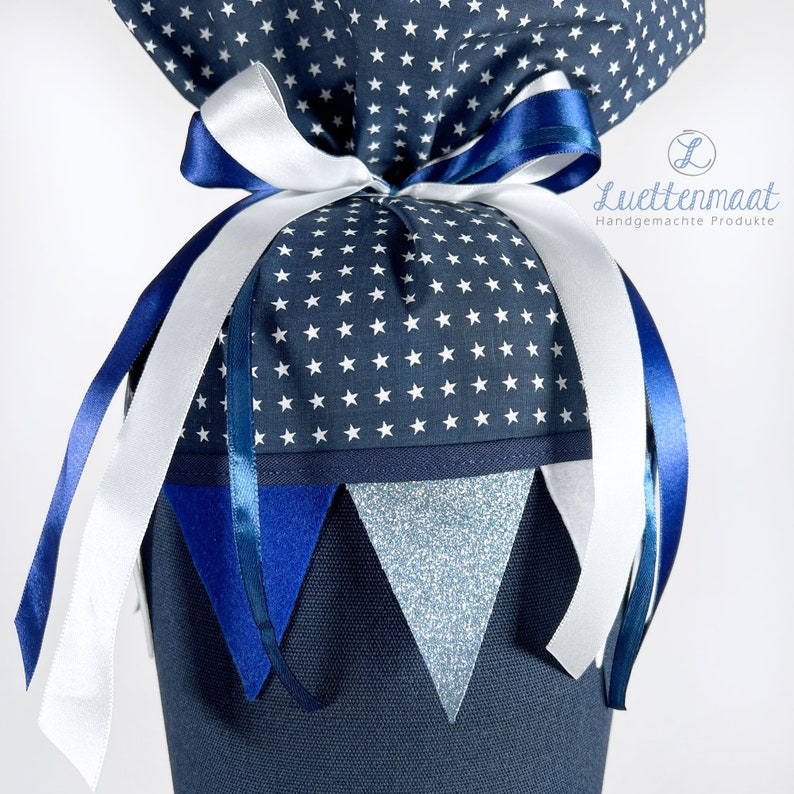 May include: A blue and white fabric gift bag with a white star pattern. The bag is tied with a white ribbon and blue ribbon. The bag has a blue and white fabric trim and a blue and silver glitter fabric trim. The bag has a tag that says "Luetjenmaat Handgemachte Produkte".