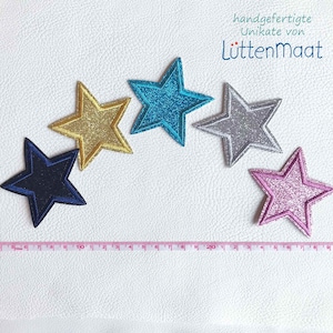 Könnte beinhalten: Fünf glitzernde sternförmige Patches in verschiedenen Farben: Gold, Türkis, Silber, Marineblau und Pink. Die Patches sind in einem Halbkreis auf einer weißen Oberfläche angeordnet. Der Text "handgefertigte Unikate von Luttenmaat" ist oben im Bild sichtbar.