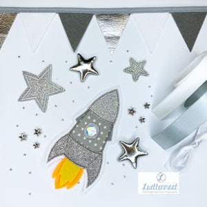 Könnte beinhalten: Eine silberne Glitzerrakete mit einer gelben Flamme, umgeben von silbernen Glitzersternen auf weißem Hintergrund. Der Text "Luettenmaat Handgemaakte Produkste" befindet sich in der unteren rechten Ecke des Bildes.