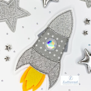 Könnte beinhalten: Eine silberne Glitzerrakete mit einer gelben Flamme, umgeben von silbernen Glitzersternen. Die Rakete hat ein Muster aus weißen Sternen und einen silbernen, schillernden Kreis in der Mitte.