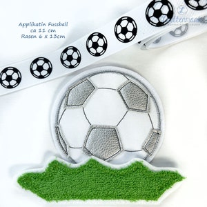 Könnte beinhalten: Ein weißes Fußball-Applikation mit silbernen Akzenten, auf einer grünen, grasartigen Basis. Ein weißes Band mit schwarzen Fußballgrafiken befindet sich darüber. Der Text auf dem Bild lautet "Applikatin Fussball ca 11 cm Rasen 6 x 13cm."