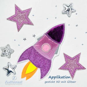 Könnte beinhalten: Ein lila Glitzer-Raketen-Applikation mit gelben Flammen, umgeben von Glitzer- und silbernen Stern-Applikationen. Die Rakete hat einen rosa Kreis in der Mitte. Der Text "Applikation gestickt 3D mit Glitzer" befindet sich unten rechts.