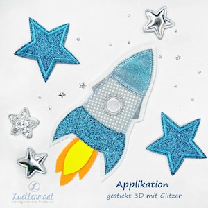 Könnte beinhalten: Ein skurriles Applikationsdesign mit einer blau glitzernden Rakete mit gelben und orangenen Flammen, umgeben von glitzernden blauen und silbernen Sternen. Das Design enthält den Text "Applikation gestickt 3D mit Glitzer".