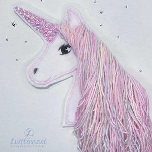 Könnte beinhalten: Weißer Einhornkopf mit rosa Glitzerhorn und rosa und pastellfarbener Garnmähne. Das Einhorn hat ein schwarzes Auge und ist mit einer rosa Kontur bestickt. Der Hintergrund ist weiß mit kleinen silbernen Sternakzenten.