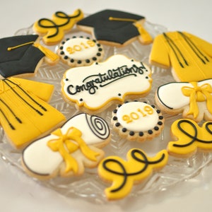 May include: A plate of decorated sugar cookies with a graduation theme. The cookies are decorated with black and yellow icing and feature images of graduation caps, diplomas, and the words "Congratulations!" and "2018" and "2019".