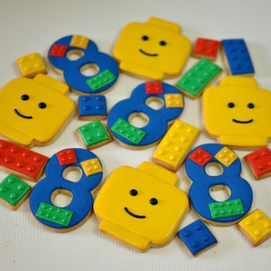 May include: A collection of colorful sugar cookies decorated with Lego-themed designs. The cookies are shaped like Lego bricks, numbers, and minifigures. The cookies are decorated with yellow, blue, red, and green icing.